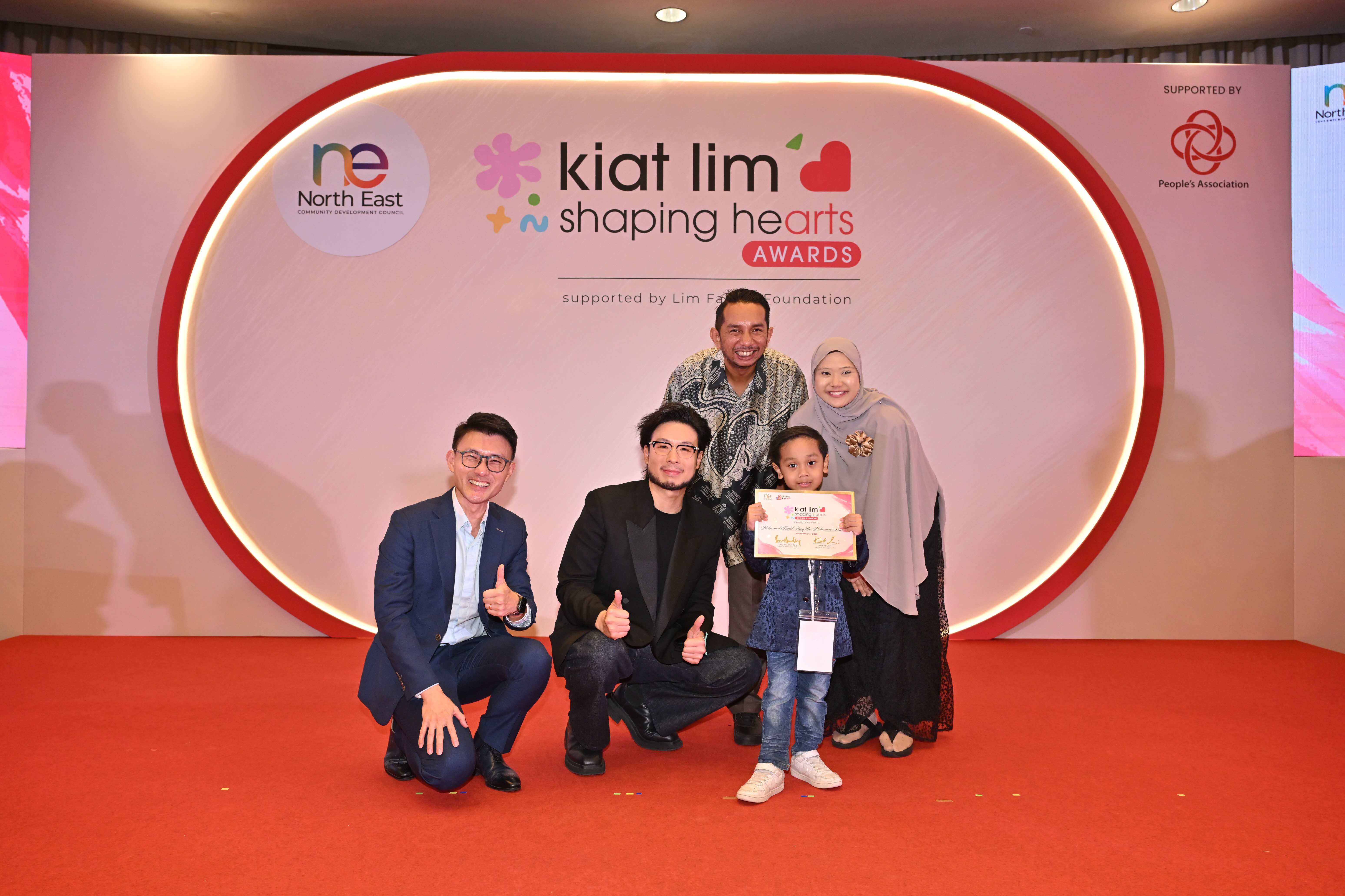 Award recipient standing on stage with the presenter, holding their certificate and posing for a group photo during the award ceremony, with a backdrop reading ‘Kiat Lim-Shaping Hearts Awards'.